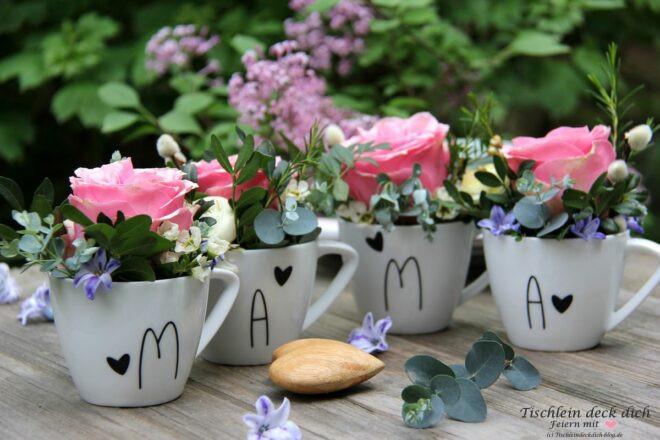 Vier Blumengestecke mit Rosen in weißen Kaffeetassen mit dem Schriftzug Mama in schwarz mit schwarzen Herzchen